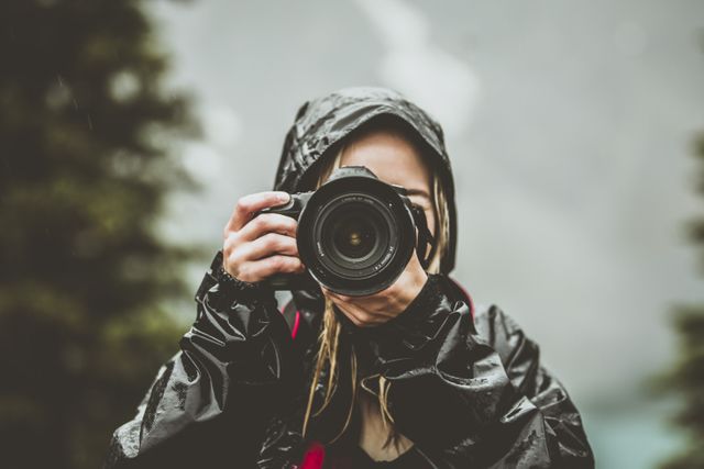 Woman in rain gear using professional camera outdoors, surrounded by nature, possibly rainy forest setting. Suitable for articles, blogs, or promotional materials related to photography, outdoor adventures, nature excursions, rainy weather gear, and capturing moments in challenging conditions.