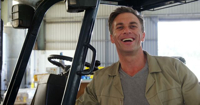 Man smiling while operating forklift in warehouse environment. Suitable for use in content related to industrial work, logistics businesses, occupational safety, and workplace positivity.
