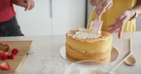 Home bakers frosting layered vanilla sponge cake with strawberries, cozy kitchen baking
