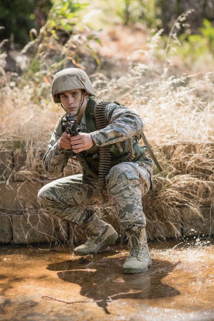 Soldier crouching by water aiming rifle during military training, representing readiness and tactical skill. Ideal for use in articles about military training, promotional materials for armed forces, tactical gear advertisements, or illustrating concepts of defense and discipline.