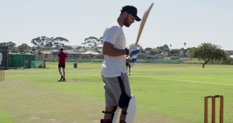 Cricketer walking off pitch holding bat wearing pads gloves and cap after practice session