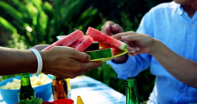 Group enjoying a summer picnic in a natural outdoor setting, with hands sharing fresh watermelon slices. Ideal for use in promotions for summer activities, outdoor gatherings, healthy eating, and family or friends reunions.