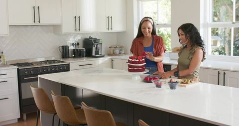 Multicultural women decorating red velvet cake with fresh berries on marble kitchen island