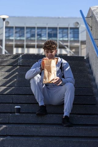 Teen male sitting on urban steps enjoying takeaway lunch and coffee, casual tracksuit