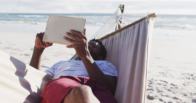 African american man using digital tablet while lying on a hammock at the beach. travel vacation lifestyle concept