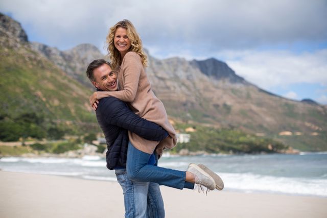 Mature couple enjoying time together on beach with mountain in background. Perfect for use in travel promotions, romantic getaway advertisements, or content related to relationships and lifestyle.