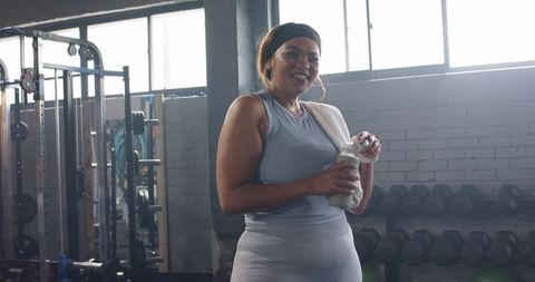 Smiling mid-adult woman holding water bottle and towel beside weight rack in backlit gym