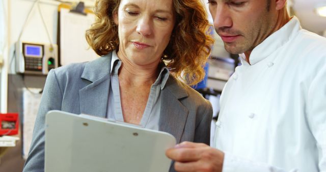 Chef in white coat collaborating with businesswoman in suit, discussing menu items or strategies using clipboard in restaurant kitchen. Ideal for themes of professional teamwork, culinary business, restaurant management, planning and collaboration.