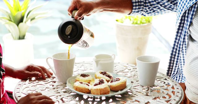 A close-up view of a couple serving coffee on a beautifully decorated patio table with fresh scones. Ideal for use in hospitality, food and beverage promotions, lifestyle blogs, and summer relaxation themes.