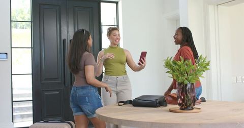 Diverse female friends greeting in sunlit modern foyer showing red smartphone