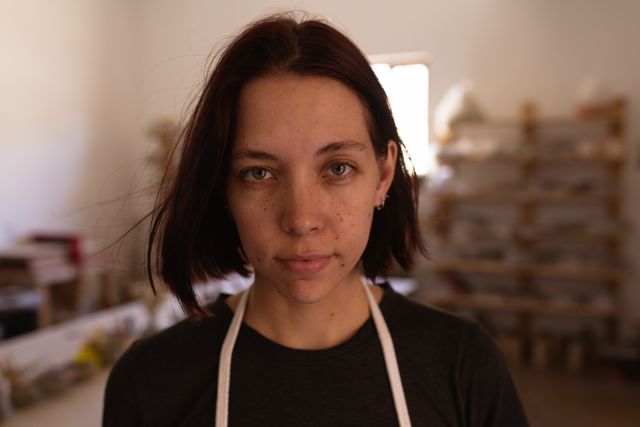 Portrait close up of a Caucasian female potter with dark red hair in a bob hairstyle, looking to camera and smiling in a pottery studio, with equipment in the background, wearing white apron.