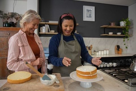 Diverse women frosting two-layer sponge cake on stand in cozy home kitchen