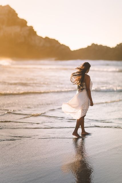 This image of a woman walking along the beach at sunset, with her flowing white dress reflecting in the wet sand, conveys a sense of calm and tranquility. It is perfect for use in travel blogs, vacation advertisements, lifestyle magazines, personal wellness articles, and social media campaigns promoting relaxation and seaside destinations.