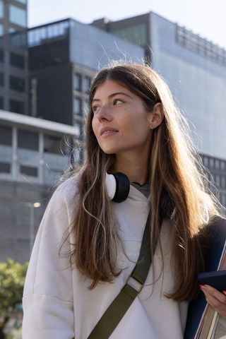 Young woman in city wearing hoodie and headphones backlit holding skateboard and phone