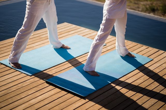 Low section of couple practicing yoga on blue mats by poolside on a sunny day. Ideal for use in wellness, fitness, and lifestyle content. Perfect for promoting outdoor activities, healthy living, and relaxation techniques.