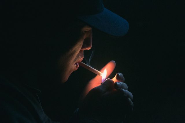 Person holding lit cigarette with lighter in dim setting. Emphasizes passion of lighting, inhaling smoke. Suitable for themes around smoking risks, addiction, solitude, late-night thoughts, evocative imagery in nighttime environment.