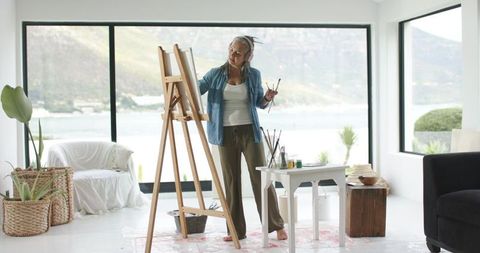 Mature woman painting at easel in sunlit coastal studio with bay windows and natural light