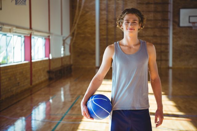 Young man standing on hardwood floor in gymnasium, holding basketball and smiling. Ideal for use in sports, fitness, and youth activity promotions, as well as educational and recreational content.