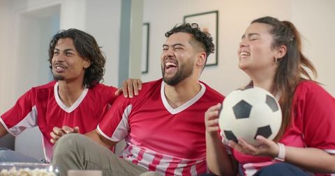 Friends cheering during soccer match at home wearing red jerseys holding soccer ball