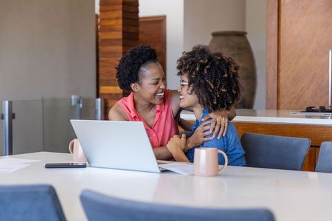 Friends embracing while working from home at kitchen table with laptop and mugs