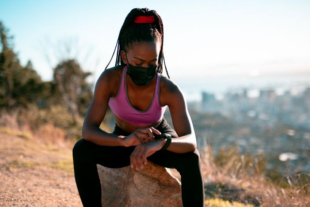 This image depicts a fit African American woman wearing a face mask and sportswear, resting on a rock while checking her smartwatch in a countryside setting. Ideal for use in articles or advertisements related to fitness, outdoor activities, health and wellness, technology in fitness, and maintaining an active lifestyle during the COVID-19 pandemic.