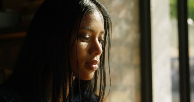 Biracial woman with long black hair looking out window. She has light brown skin, wearing minimal makeup, and thoughtful expression