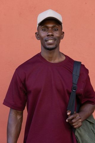 Smiling man wearing beige cap and burgundy shirt carrying olive satchel, terracotta wall