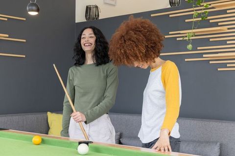 Young diverse women playing pool in modern recreation lounge holding cue laughing