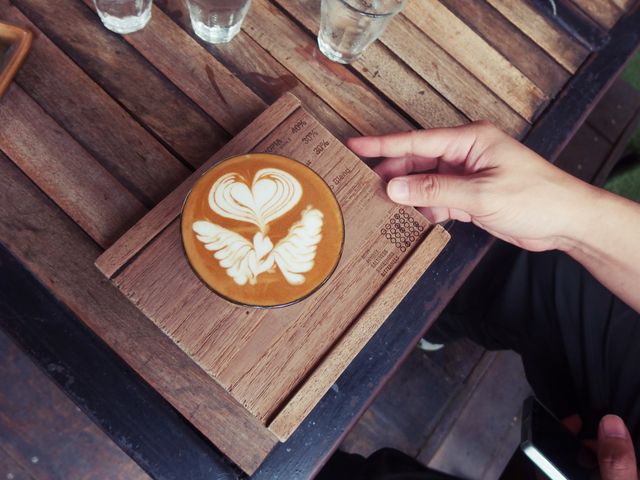 Hand holding cup of latte with intricate latte art on top, set on rustic wooden table. Perfect for illustrating relaxed cafe scenes, enjoyment of warm beverages, coffee culture, and craftsmanship of baristas. Useful for blogs, social media posts, and articles about coffee, cafes, and lifestyle.
