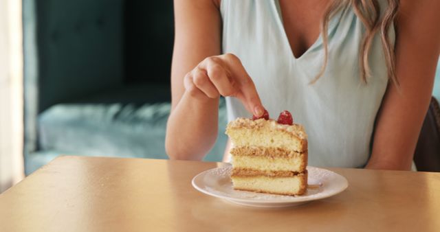 Woman touching top of a vanilla cake slice while sitting in a cozy cafe. Suitable for use in dessert or bakery advertisements, articles about cafe culture, or as a visual in culinary blogs.