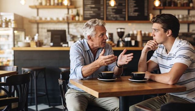 Older man mentoring young man over coffee in cozy rustic cafe discussing life and career