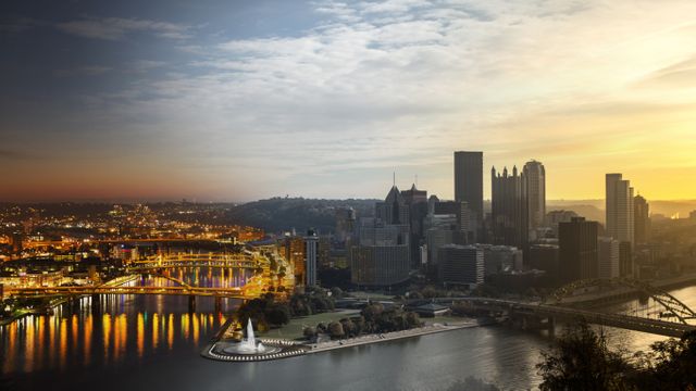 City skyline of Pittsburgh at dusk featuring Point State Park with lit buildings and bridges reflecting on rivers. Perfect for travel guides, tourism promotion, or illustrations showcasing Pittsburgh's urban beauty.