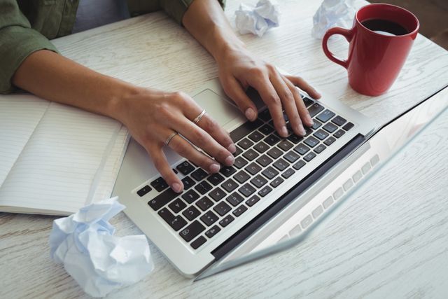 Businesswoman typing on laptop at desk with coffee and crumpled papers. Ideal for illustrating work environment, productivity, office life, business tasks, and remote work scenarios.