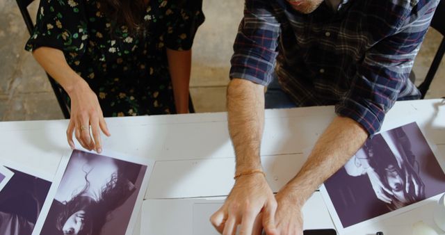 Two individuals, a Caucasian woman and a Caucasian man, are engaged in examining photographs spread out on a table, with copy space. Their collaborative effort suggests a creative or editorial process, reviewing photographic work for selection or critique.