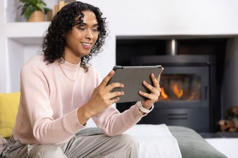 Non-binary adult using tablet in cozy modern living room with burning fireplace