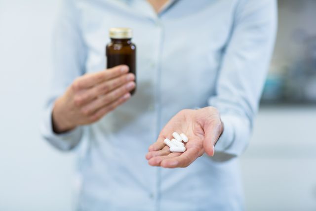 This image depicts a pharmacist holding a medicine bottle and several pills in their hand, likely in a pharmacy setting. It can be used for articles or advertisements related to healthcare, medication, pharmaceutical services, or pharmacy operations. It is suitable for illustrating concepts of prescription medication, healthcare professionals, and medical treatments.
