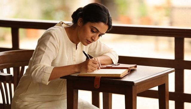 Indian woman writing in leather journal on wooden veranda table in cream kurta, reflective