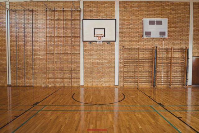 Empty high school basketball court with a polished wooden floor and a basketball hoop mounted on a brick wall. Ideal for use in educational materials, sports facility promotions, and articles about school sports programs.