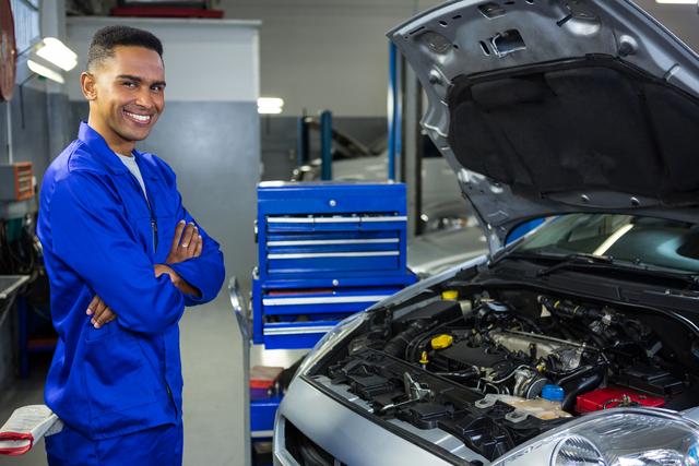 Mechanic standing confidently in front of an open car hood, with arms crossed in a professional auto repair garage. Ideal for use in promotions for automotive services, car maintenance businesses, mechanical engineering content, and vocational training programs.