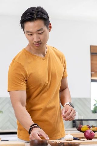 Asian man chopping garlic on cutting board in bright modern kitchen wearing mustard tee
