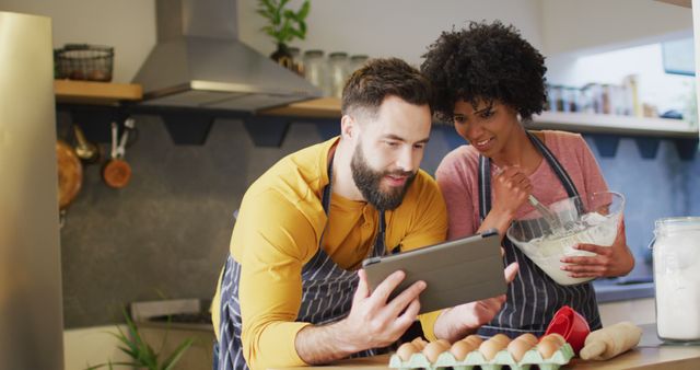 Image of happy diverse couple in aprons using tablet, baking together in kitchen, with copy space. Happiness, communication, inclusivity, free time, togetherness and domestic life.