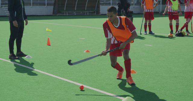 A young hockey player, dressed in bright orange training attire, focuses on handling the hockey stick and ball in an outdoor field setting. Teammates in action and a coach are in the background, suggesting a training session or team practice. This photo is ideal for use in promotions related to youth sports, team-building activities, school gym classes, and articles about the importance of exercise and sportsmanship.