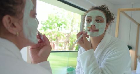 African american woman applying green facial mask in white robe at vanity mirror