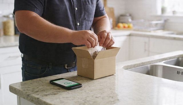 Man Unpacking Cardboard Box on Kitchen Counter While Smartphone Showing Delivery Confirmation