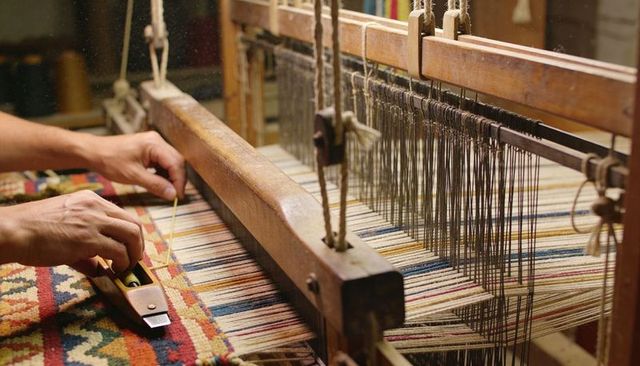 Artisan weaving traditional geometric rug on vintage wooden floor loom with shuttle and hands
