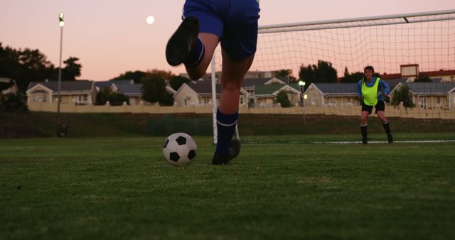Soccer player preparing to kick ball towards goal during twilight with goalie in position. Can be used for illustrating aspects of sports action, training intensity, passion for soccer, and dusk activities.