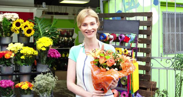 A cheerful female florist stands in her flower shop, holding a colorful bouquet. She is wearing an apron and looks happy while presenting the beautifully arranged flowers. The shop has various flowers, plants, and floral supplies displayed. This image can be used for advertising local businesses, websites about entrepreneurship or floristry, or articles on small business success.