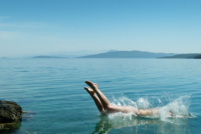 Person diving into calm, clear lake with splash, surrounded by mountains and clear blue sky. Ideal for use in travel blogs, summer vacation advertisements, and outdoor adventure promotions.