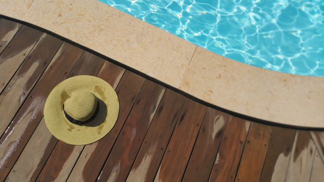 Straw hat lying on wooden deck next to a brightly lit swimming pool with clear blue water. Perfect for illustrating outdoor relaxation, summer vacations, or poolside fun. Useful for travel magazines, vacation brochures, and advertisements for summer essentials.