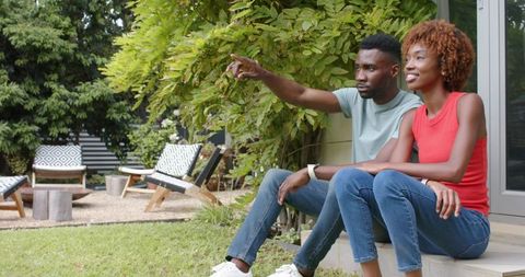 Young black couple relaxing on front steps pointing toward backyard lounge and greenery